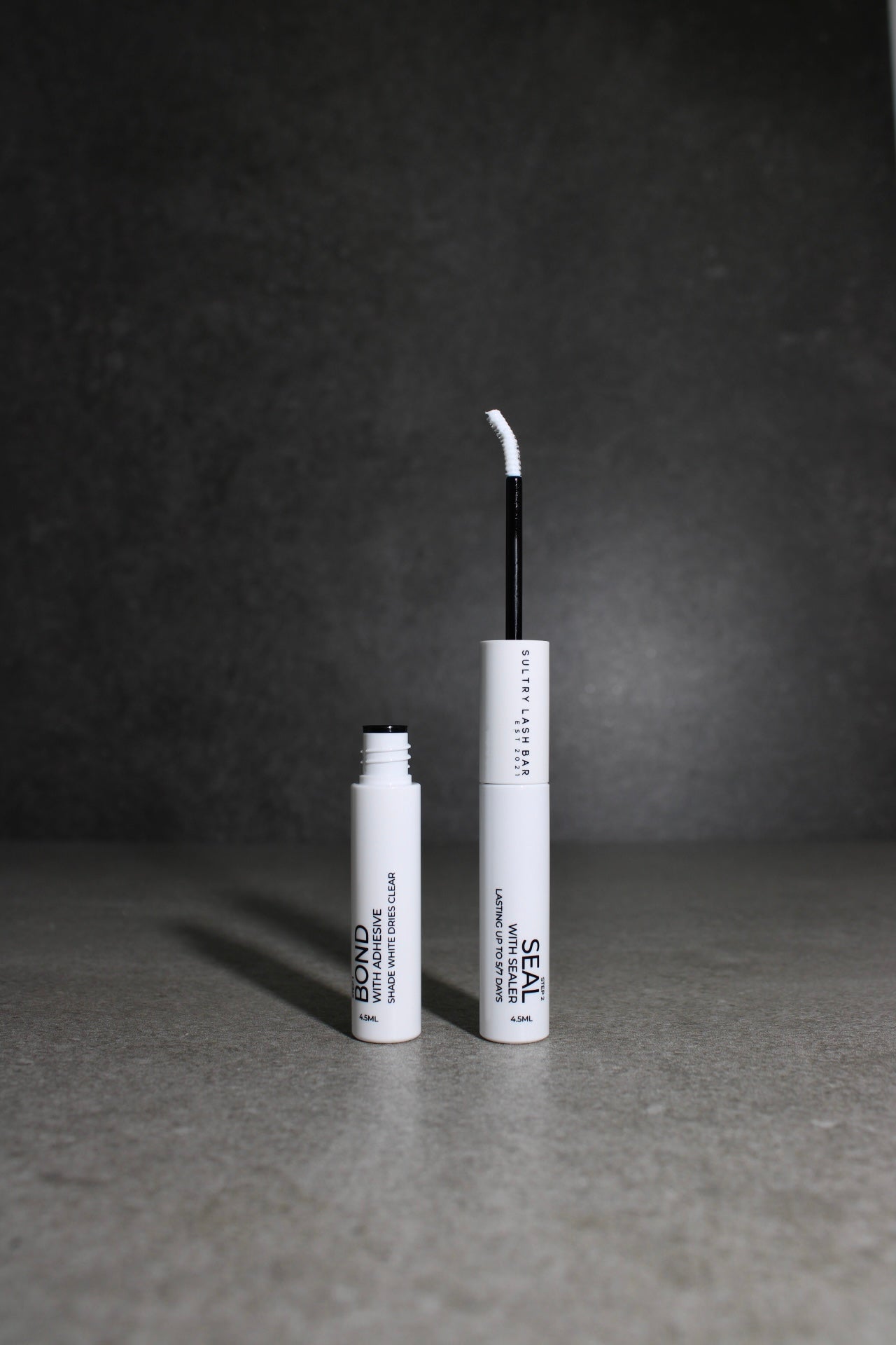 Two white cosmetic bottles with black text on a gray surface and dark background