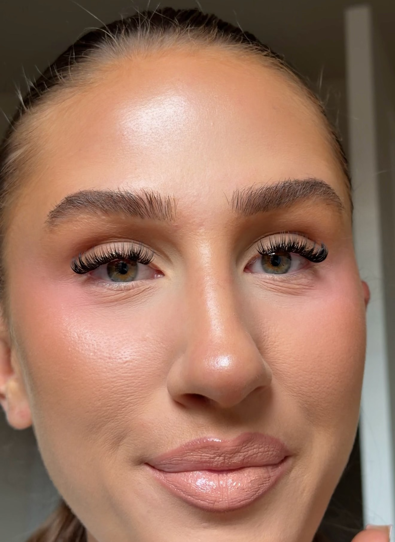 Close-up of a woman's face showcasing makeup, featuring defined eyebrows, long eyelashes, and a glossy nude lip, with her hair pulled back.